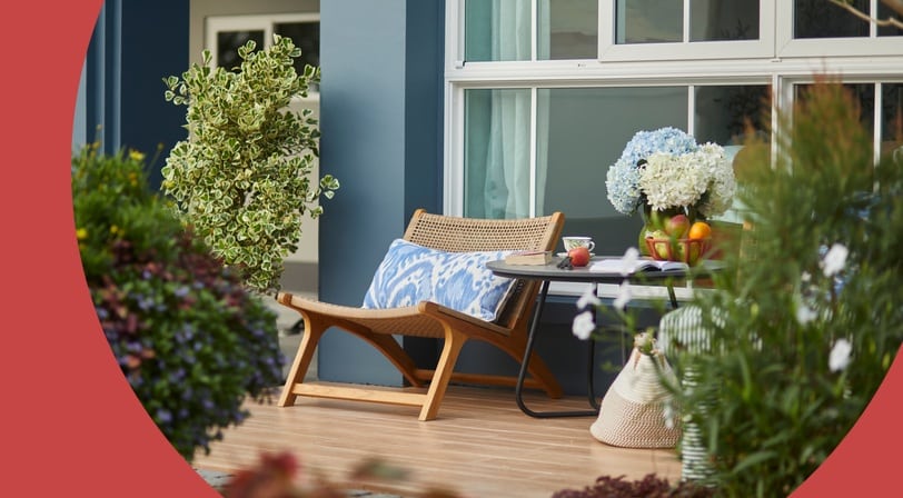 Woven lounge chair and small table with flowers and fruit on a front porch surrounded by blooming garden beds.