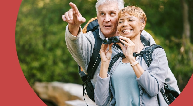 An older couple pauses on a wooded hiking trail, one holding binoculars while the other points toward something in the distance.