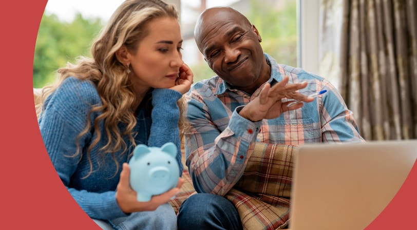 A couple sits together on a sofa reviewing finances on a laptop while holding a blue piggy bank.