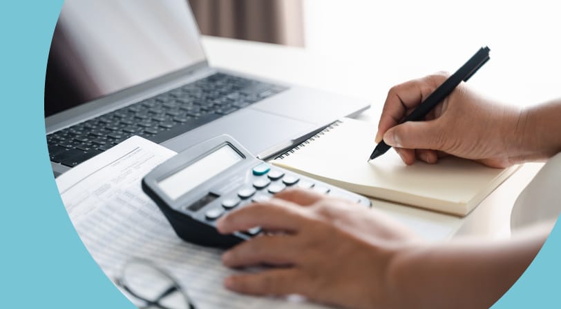 Close-up of hands using a calculator and writing notes beside tax paperwork, a laptop, and reading glasses on a white desk.