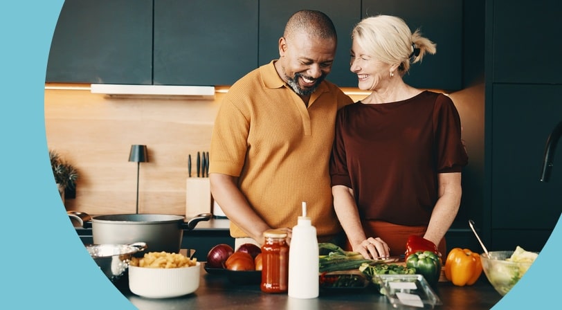 An older couple laughs together while preparing a meal of fresh vegetables and pasta in a modern kitchen.