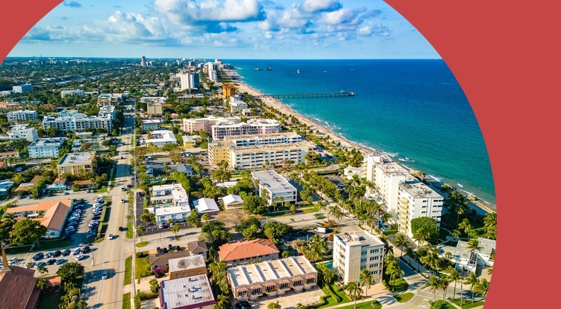 Aerial view of Deerfield Beach, Florida with low-rise condominiums, palm-lined streets, a sandy beach, and a fishing pier.