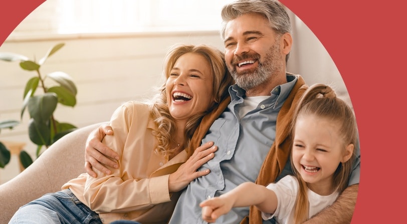A couple and their young daughter laugh together on a sofa in a sunlit living room.