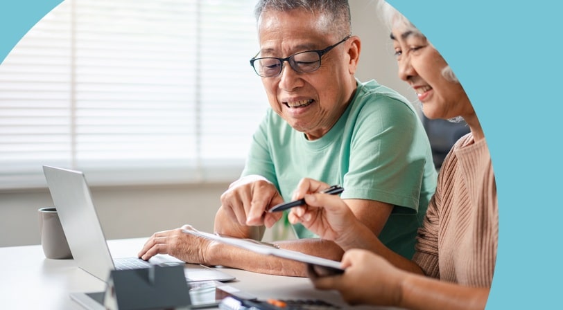 A senior couple sit together at a table reviewing documents on a laptop with a calculator and small model house nearby.