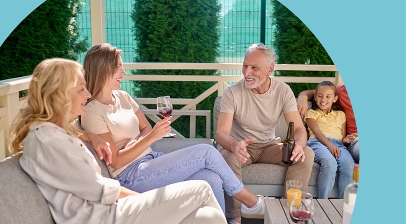 A 55+ couple and their adult children and granddaughter laughing together over drinks on a covered patio.