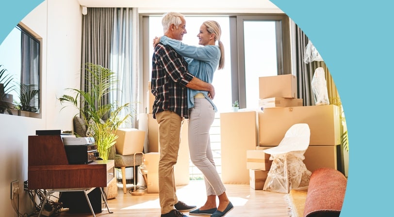 A mature couple embraces in the living room of their new home, surrounded by moving boxes and unwrapped furniture.