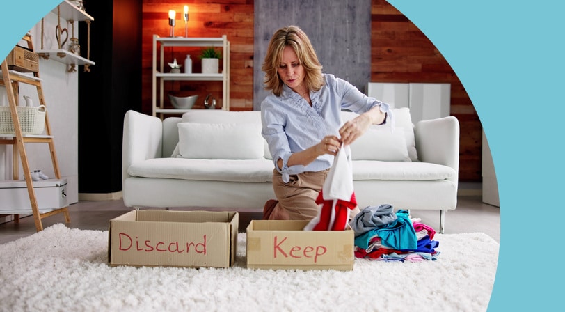 A woman kneels on a living room rug sorting folded clothing into cardboard boxes labeled "Keep" and "Discard."