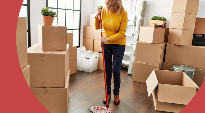 A woman mops the hardwood floor of a new home surrounded by stacked moving boxes and a potted plant.
