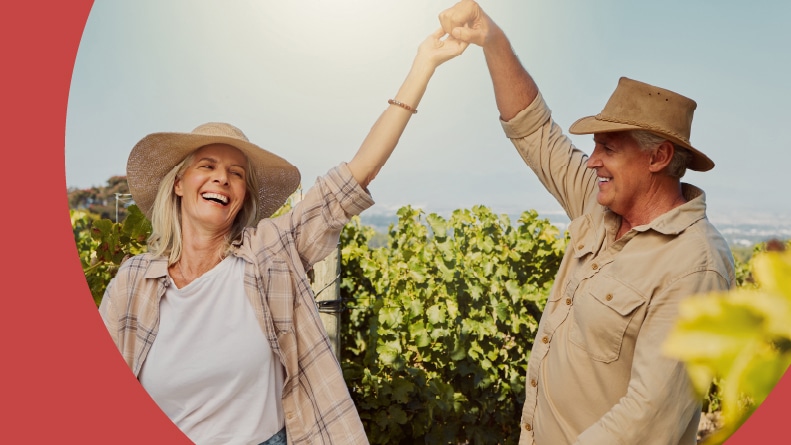 An older couple in sun hats dances joyfully between rows of grapevines at a sunny vineyard.