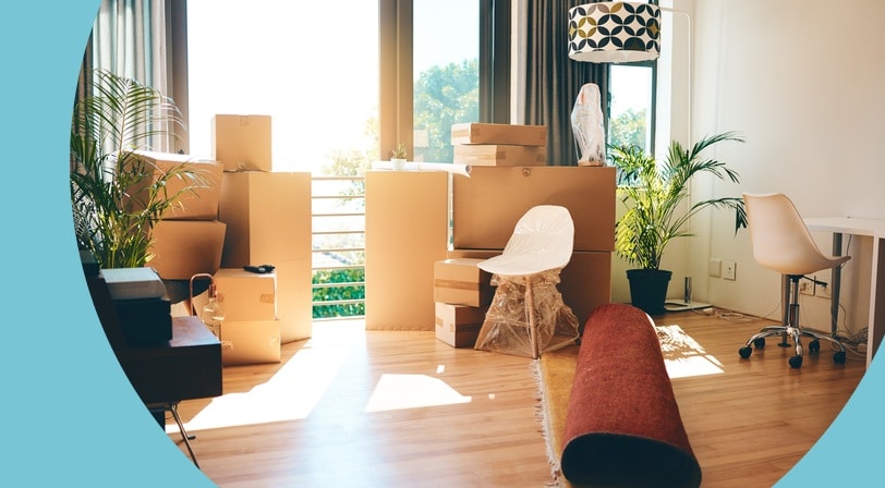 Stacked cardboard moving boxes, wrapped furniture, and a rolled rug fill a sunlit living room on moving day.