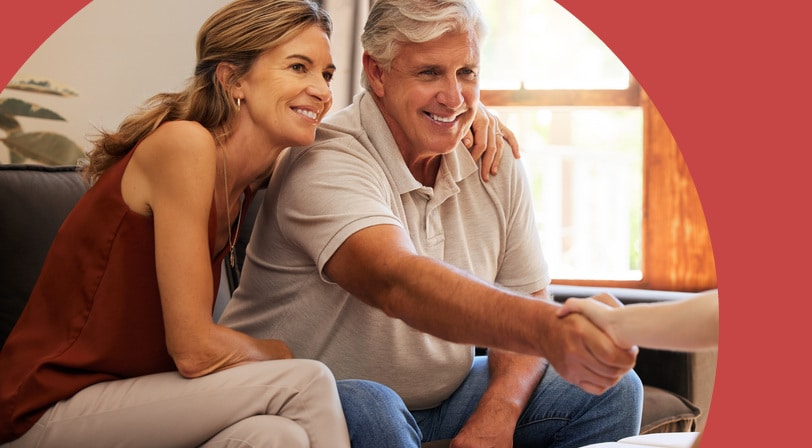 A smiling mature couple sits on a sofa in their living room shaking hands with a real estate agent across a table of paperwork.