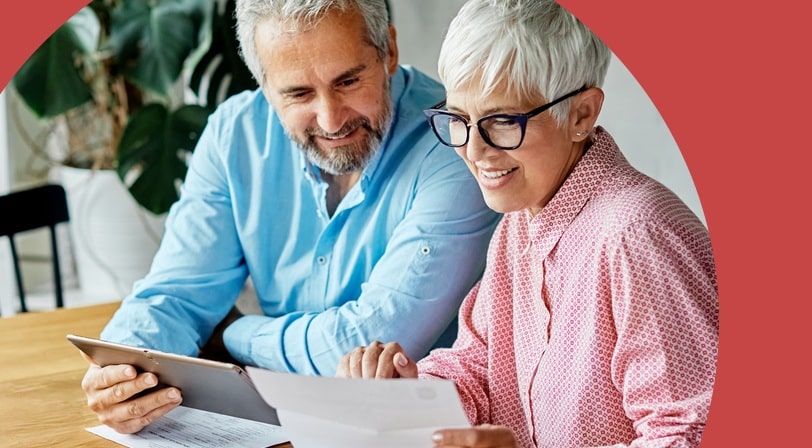 An older couple sit at a dining table reviewing printed paperwork together, one holding a tablet.