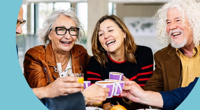 A group of 55+ friends laughing and toasting with coffee and juice at a cafe.