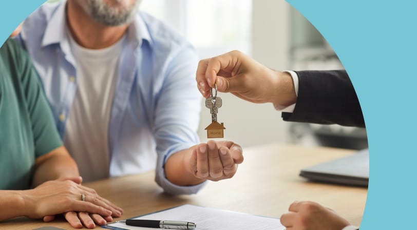 A real estate agent hands a set of keys with a house-shaped keychain to a smiling couple seated at a desk with a signed contract.