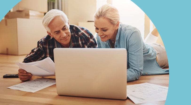 A mature couple lies on the hardwood floor of their new home, smiling as they look at a laptop and floor plans with moving boxes behind them.