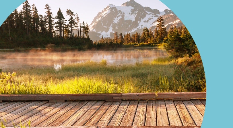 Wooden boardwalk beside a misty alpine lake with snow-capped Mount Shuksan rising behind evergreen trees.