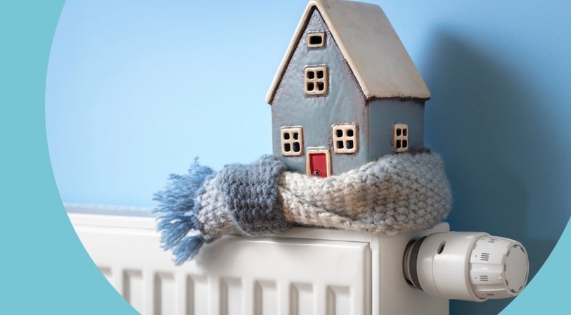 Miniature ceramic house wrapped in a knitted scarf sitting on top of a white radiator against a blue wall.