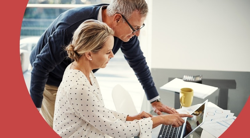 A 55+ couple reviewing a laptop together at a desk, with papers and a mug nearby.