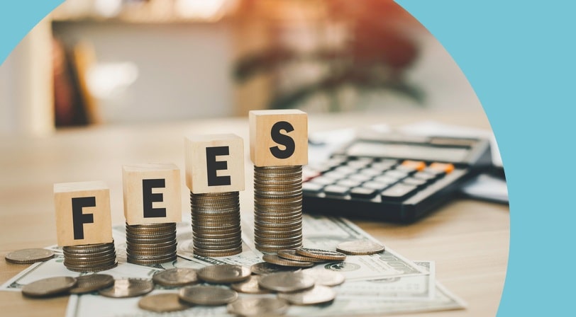 Wooden blocks spelling "FEES" resting on rising stacks of coins beside a calculator and cash on a desk.