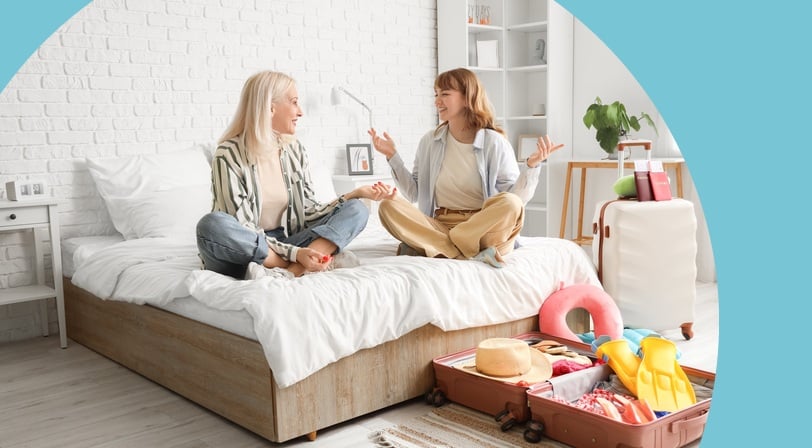 A mother and adult daughter sit cross-legged on a bed chatting while packing open suitcases for a trip.