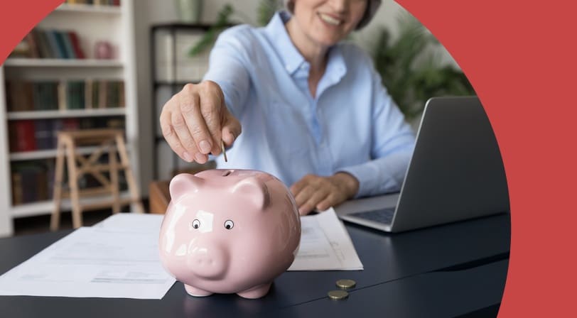 A smiling 55+ woman drops a coin into a pink piggy bank at her desk, laptop and bills nearby.