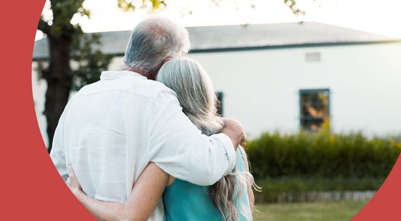 An older couple stands with arms around each other, looking toward a white farmhouse framed by a large tree and garden hedges.
