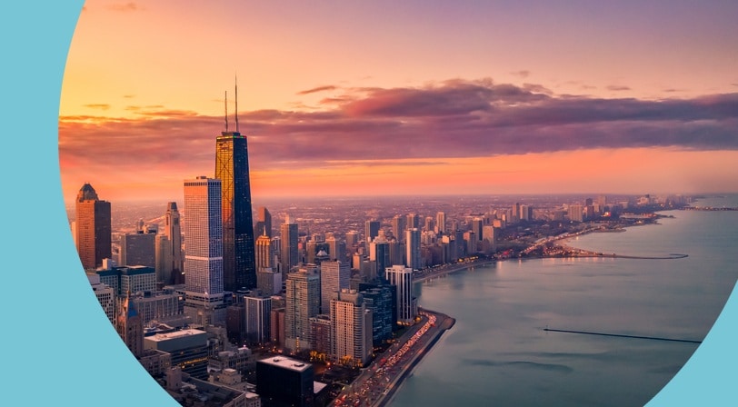 An aerial view of a city skyline along a lakefront at sunset, with a prominent skyscraper lit by golden light.