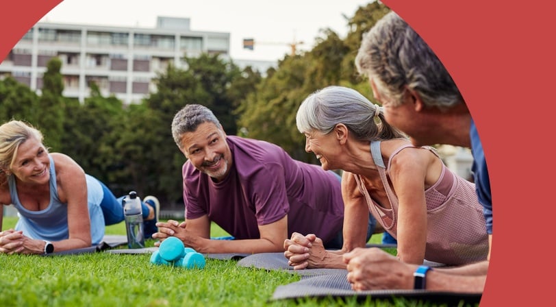 A group of four smiling adults hold plank positions on yoga mats in a grassy park, with teal dumbbells and a water bottle nearby.