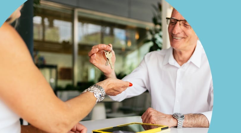 A smiling man in a white shirt hands over a set of keys to a woman across a table with a yellow-cased tablet.