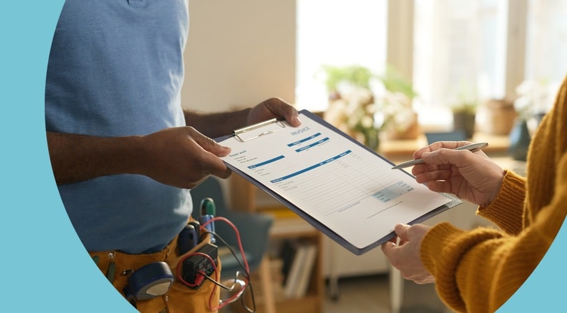 A contractor wearing a tool belt handing a clipboard invoice to a customer, who points at the total with a pen.