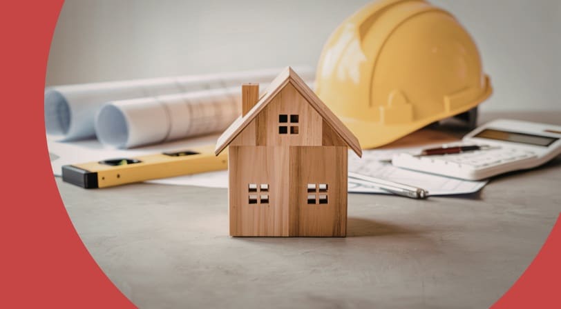 A small wooden model house on a desk beside rolled blueprints, a yellow hard hat, a level, and a calculator.