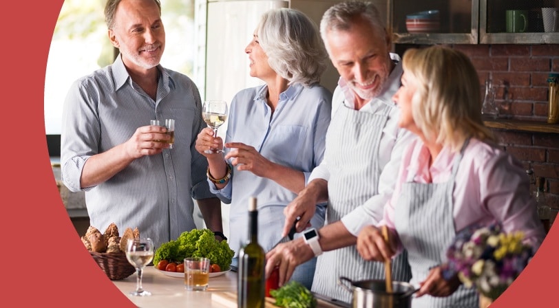 Two older couples laugh and share drinks while preparing a meal together in a rustic kitchen.