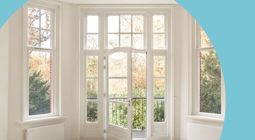 A sunlit room with white french doors open to a balcony, flanked by tall windows overlooking autumn trees.
