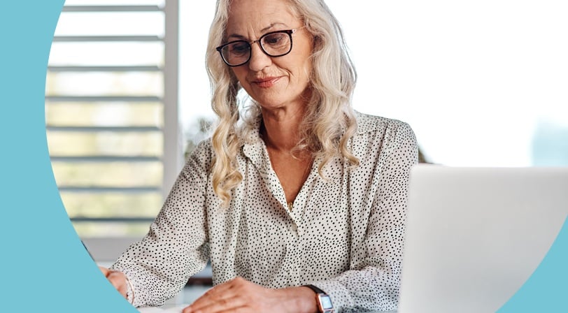 An older woman wearing glasses writes in a notebook at a desk while working on her laptop near a sunny window.