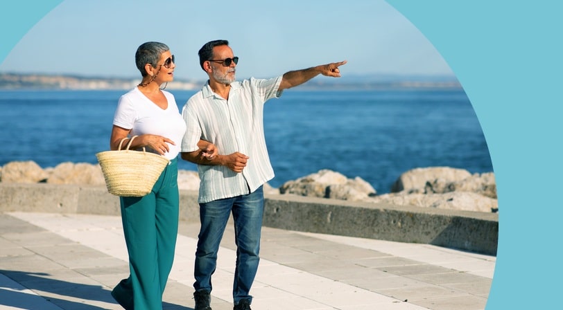 A smiling couple walks arm-in-arm along a waterfront promenade, one pointing out toward the sea.