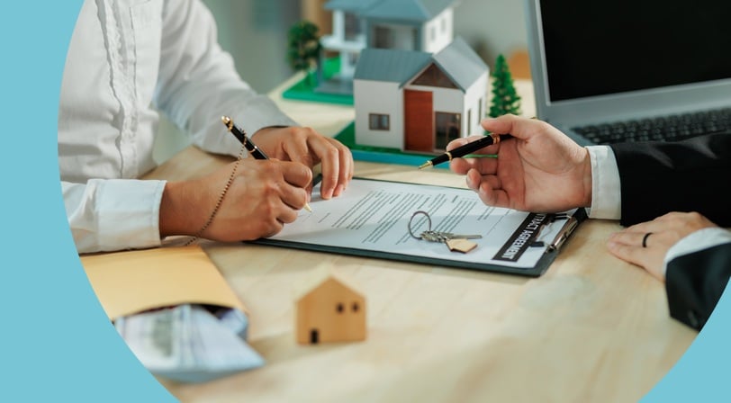 Two people seated at a desk reviewing and signing a home purchase agreement, with house keys and a model home nearby.