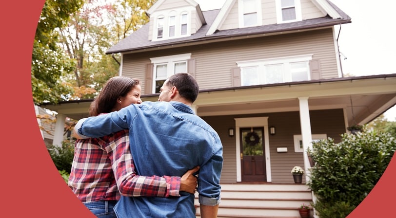 A 55+ couple embraces from behind, admiring a two-story craftsman-style house with a wraparound porch.