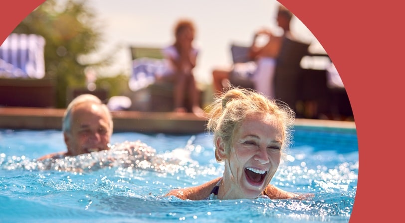A 55+ woman laughs while swimming in a pool alongside her partner, with blurred figures relaxing poolside in the background.