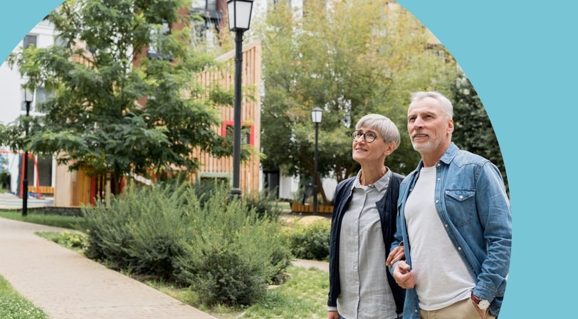 A 55+ couple walks arm-in-arm along a paved path through a landscaped courtyard surrounded by apartment buildings.
