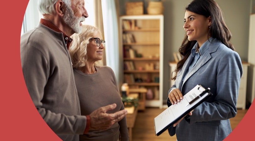 An 55+ couple speaks with a real estate agent holding a clipboard inside a home with a bookshelf in the background.