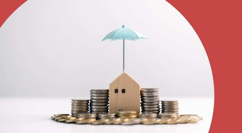 A small wooden house model shaded by a miniature umbrella and surrounded by stacked coins, symbolizing financial protection and retirement planning.