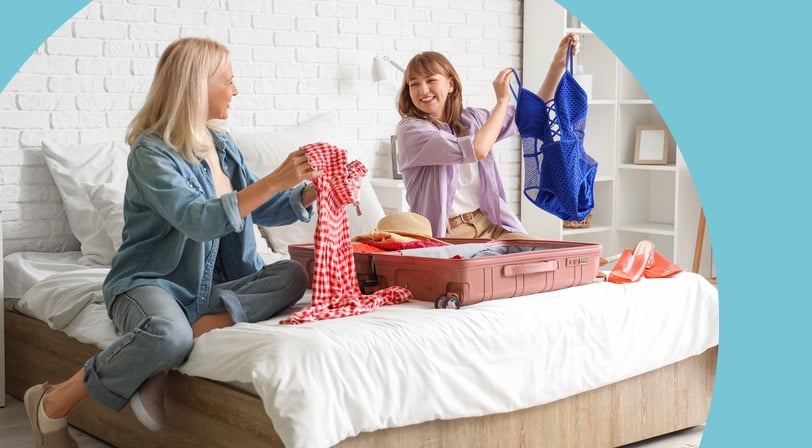 Two women laughing while packing an open suitcase on a bed.