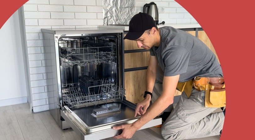 A technician in a tool belt kneeling in a bright kitchen to install a stainless steel dishwasher.