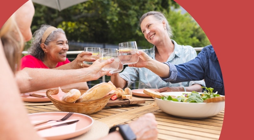Four 55+ women toast with glasses of lemon water at an outdoor patio table set with bread and salad.
