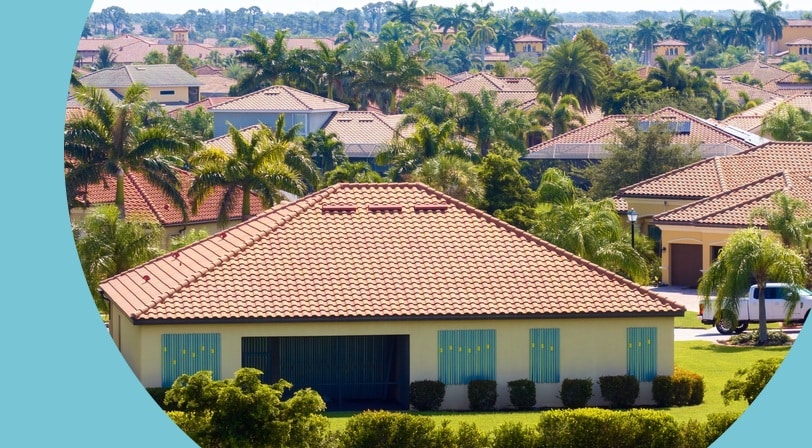 Single-story home with a terracotta tile roof and hurricane shutters surrounded by palm trees in a Florida neighborhood.