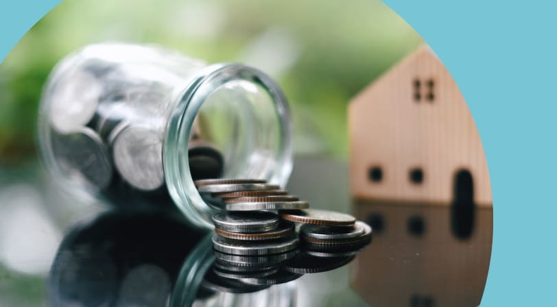 A glass jar tipped on its side with coins spilling out, a small wooden model house in the background.