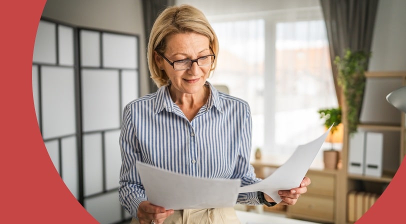 A 55+ woman in glasses reading through papers while standing in a bright home office.