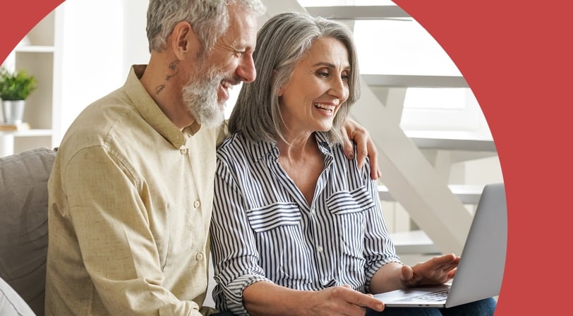 A smiling 55+ couple sitting close together on a couch, looking at a laptop screen.
