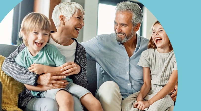 Grandparents laugh with their two young grandchildren while sitting together on a living room sofa.