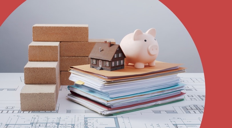 A pink piggy bank and miniature house on a stack of file folders beside bricks and construction blueprints.